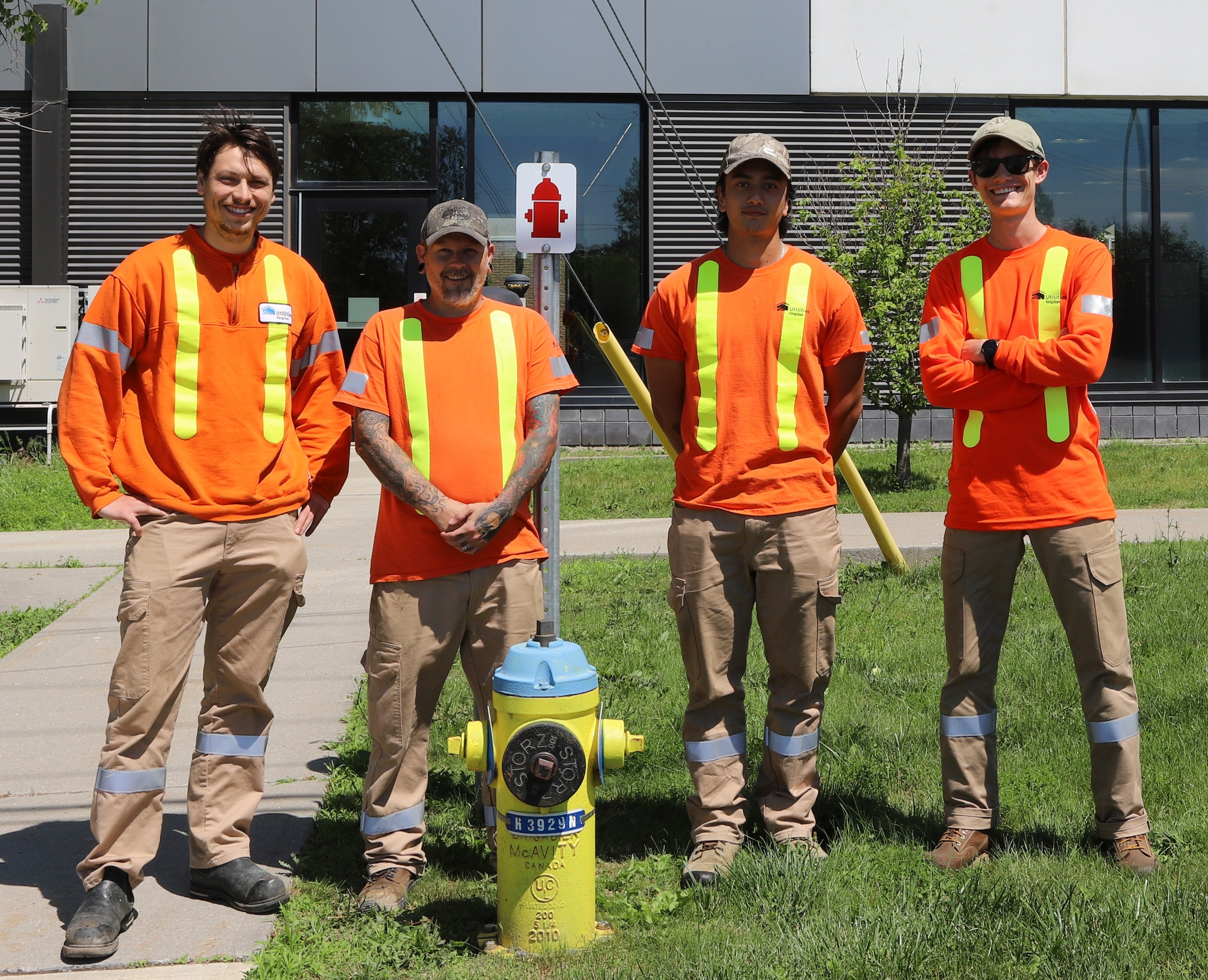 Four Utilities Kingston workers in orange high-visibility shirts stand on grass near a yellow fire hydrant in front of a modern building.